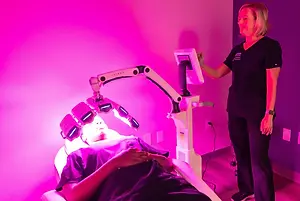 A woman is lying on a bed under a pink light, and a woman is standing beside her holding a monitor. The woman lying down is wearing a purple shirt, while the woman standing is wearing a black shirt with a logo on it. There is a pink wall with a power outlet and a plug in the background.