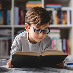 A boy wearing glasses is reading a book on his bed.