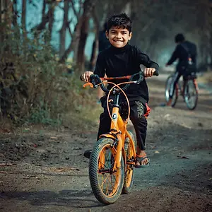 The boy is riding a bicycle on the road, he is smiling while riding the bicycle and holding the handle of the bicycle, and there is another person riding a bicycle behind him. The road is made of soil, and there are some plants on the side of the road. In the distance, there are some trees and a blurry view of a person. The shadow of the boy is falling on the road.