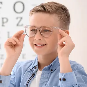 A young boy wearing glasses is smiling and posing for a photo in front of an eye chart.