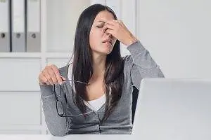 A woman sitting in front of a laptop with her eyes closed and holding a pair of glasses