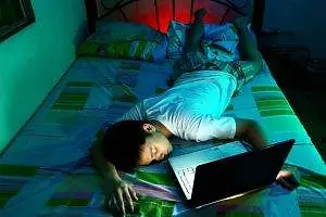 A man is sleeping on a bed while using a laptop, with his eyes closed and head resting on a pillow, and a red light illuminating the room