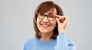 A smiling woman wearing a blue sweater and glasses, adjusting her glasses with her right hand.