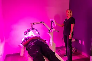 A woman is standing next to a man who is lying on a bed with a light on his face. The woman is holding a tablet in her hand, and she is probably explaining something to the man. Behind them is a wall with a pink light. There are power outlets and a stool in the room.