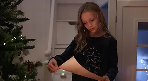 A young girl stands in front of a Christmas tree, holding a piece of paper and looking down at it with curiosity, with a staircase and a lit window in the background.