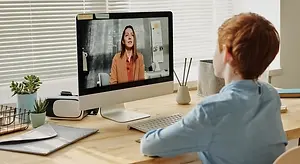 A young boy is sitting in front of a computer screen, watching a video on Zoom. He is wearing a blue shirt and appears to be focused on the video. The computer screen displays a woman giving a presentation, and the background includes a desk with a potted plant, a book, and a white keyboard. The desk is located in a room with white blinds and a white wall.