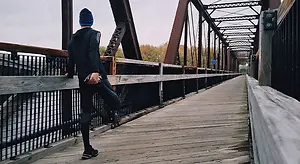 A man dressed in a black hoodie and black pants stands on a wooden bridge, leaning on the wooden railings, with a cloudy sky in the background.