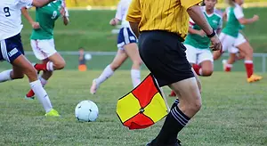 A soccer player in a yellow shirt holds a flag next to a soccer ball on a grassy field as other players run behind him wearing green jerseys and sneakers