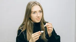 A woman with long blonde hair is holding a small white object in both hands, sitting in a room with a white wall, possibly posing for a photo.