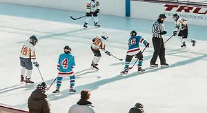 A group of hockey players are on an ice rink with their shadows cast on the ice, and some of them are wearing jerseys with numbers and helmets. The referee is wearing a striped shirt and holding a hockey stick. The ice rink has a white wall with some text written on it, and there are people sitting on the bleachers watching the game.