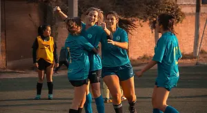 A group of female soccer players are celebrating on a field with a brick wall in the background