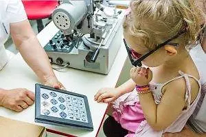 A young girl is at an optometrist’s office, sitting on a chair, wearing glasses, and holding a tablet with a grid pattern while an adult holds a tablet and adjusts a machine behind her.