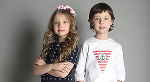 Two children posing for a photo with a gray background. A girl with curly hair wearing a blue shirt with a flower crown and a boy wearing a white shirt with an anchor design.