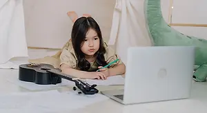 A young girl studies with her ukulele and laptop on the floor in a cozy setting.