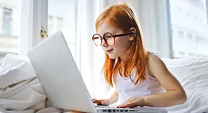 A young girl with red hair and glasses is sitting on a bed and using a laptop computer.
