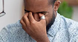 A man is holding his nose and seems to be in pain. He is wearing a ring on his right hand and a blue polka-dot shirt. Behind him is a blurry image of a plant with green leaves. On the left side is a blurry image of a white wall with a silver object on top, probably a lamp.