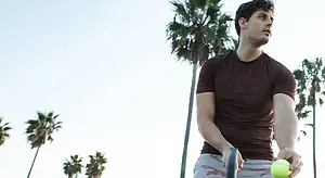 A man holding a tennis ball, dressed in a brown t-shirt and gray shorts, standing in front of a palm tree, possibly playing tennis.