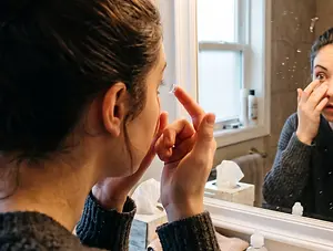 A woman in a bathroom looks at her reflection in a mirror as she applies eye drops.