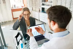 A man wearing a white lab coat and a woman wearing a gray blazer and glasses sit across from each other, the man is holding a contact lens and the woman is smiling while the man shows her something on a laptop.