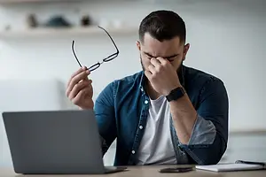 A man is sitting in front of a desk, holding a pair of glasses, and covering his face with his hand, possibly indicating discomfort or a headache. He is wearing a denim jacket and a white shirt, and there is a laptop, a book, and a cell phone on the desk. Behind him, there is a shelf with various items, including a bottle, a cup, and a box.