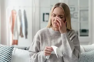 A woman is sitting on a couch holding a tissue and covering her face with her left hand while looking at something