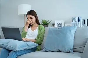 A woman sits on a couch holding a laptop while rubbing her eyes, indicating fatigue.