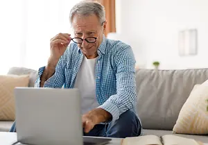 An elderly man wearing glasses and a blue checkered shirt is sitting on a couch and working on his laptop.
