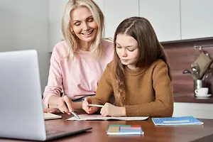 A woman and a girl are sitting in front of a desk, possibly in a kitchen, and they seem to be working on a project. The woman is smiling while looking at the girl, who is holding a pencil and a pen. The desk has a laptop, notebooks, and other items. Behind them is a countertop with a sink, a coffee cup, and a knife holder.