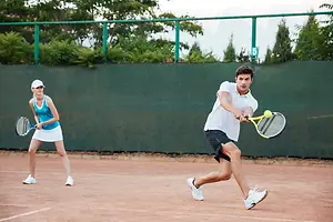 A man and a woman playing tennis on a court. The man is about to hit the ball. The woman is standing and looking at him. They are both wearing white shoes and hats.