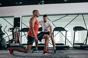 A man is performing a lunge exercise while a trainer watches closely in a gym with treadmills and weights in the background.