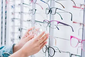A woman holding a pair of glasses in her hands in front of a wall with multiple pairs of glasses hanging on it