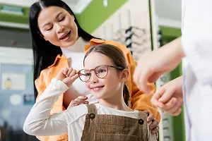 A young girl wearing glasses is being examined by an adult woman and another person in a room