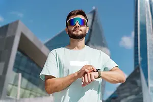 A man in sunglasses and a t-shirt is standing outside and looking at his smartwatch.