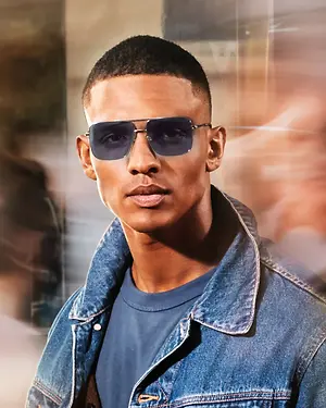 A man wearing sunglasses and a denim jacket is standing in front of a glass window. The man's face is slightly blurred.