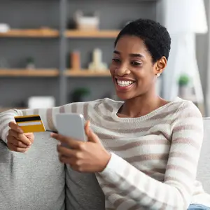 A woman with a short hairstyle smiles while holding a credit card and a cell phone in her hand.