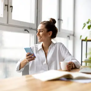 A woman sitting at a table with a cup of coffee and a book in front of her, holding a cell phone in her hand and smiling.