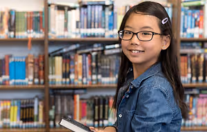 A young girl holding a book in a library wearing eye glasses