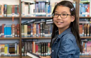 A young girl holding a book in a library wearing eye glasses