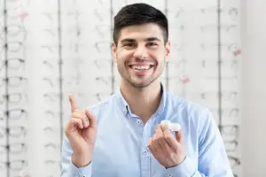 A smiling man with glasses and a blue shirt holding a white object in his hands in front of a wall of eyeglasses.