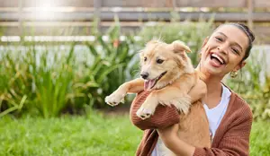 A woman holding a dog in a yard with green grass and plants in the background