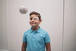A young boy is wearing an eye patch while looking up at a white ball with red letters that is floating in the air in a room.