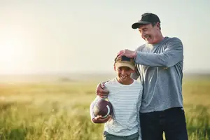 A man and a young boy are standing in a field, with the man holding the boy's head and smiling, and the boy holding a football and also smiling.