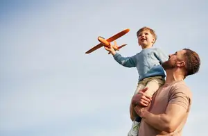 A man with a beard is holding a young boy on his shoulders and they are both looking up at a toy plane in the sky.