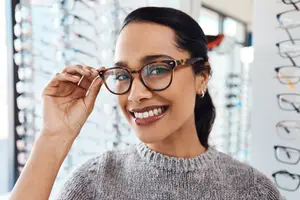 Woman in gray sweater wearing brown-framed glasses smiling and posing for a photo inside a store with glass walls displaying glasses.