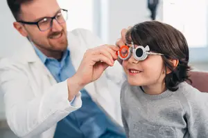A man in a white lab coat is examining a young girl's eyes with a special tool. The girl is wearing glasses and smiling at the man.