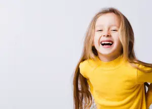 A little girl with blonde hair wearing a yellow sweater is smiling and looking at something