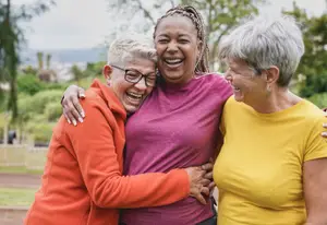 Three women standing together and laughing, two are hugging the woman in the middle and one is holding her waist