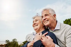 An older couple embracing and smiling in an outdoor setting with trees and a blue sky in the background