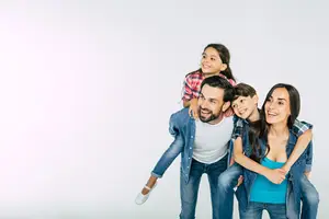 A smiling family of four, two kids on a man's shoulders and one on a woman's side, standing together for a photograph