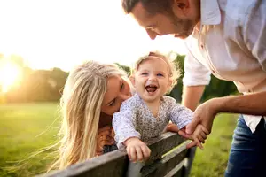 A family of three standing in a park, the father is holding the hands of the toddler while the mother is looking at the baby girl smiling